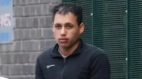 Eddie Mitchell Man in a black zipped jacket stands outside by a brick wall and metal fencing, looking toward the camera.
