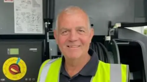 George Irwin wear high vis jackets. They stand in front of a street washing machine.