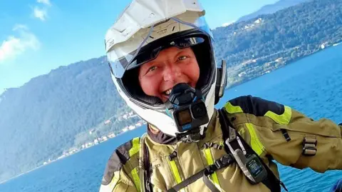 Jodie Rogers Jodie Rogers is smiling wearing a white motorbike helment, and a khaki jacket with yellow flourescent strips. Behind her is a body of water with a hilly coastline in the distance with buildings on a bright and sunny day.