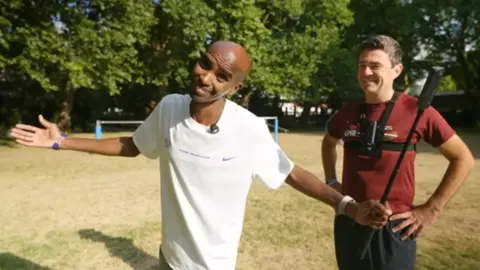 Sir Mo Farah Sir Mo Farah smiles and spreads his arms wide, as he holds a selfie stick in a park with dry, parched grass and trees. He is wearing a white Nike T-shirt. Andy Burnham is standing behind him with his hands on his hips. He is wearing a red T-shirt and jogging bottoms.
