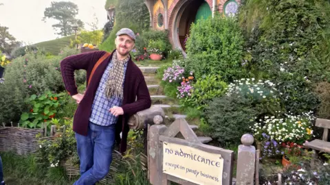 Kyle Pedley A man in a flat cap, maroon jacket, chequed shirt and jeans leaning against a gate with a garden and a round doorway behind him
