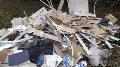 The Parks Trus A large pile of fly-tipped waste is dumped on vegetation beside a wooded area. The heap includes broken plasterboard, timber, cardboard, plastic bags, packaging, a door and other construction and household debris.