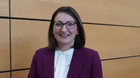 Department of Health A woman with a brunette bob smiles at the camera - she wears a purple jacket and tortoiseshell glasses