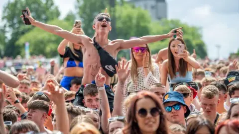 Music fans in a crowd dressed in summery and festival clothing. People are up on shoulders, including one male and three females 