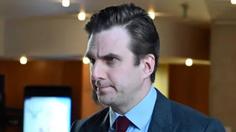 Getty Images Daniel Johnson in a grey suit, light blue shirt and red tie is photographed indoors, with blurred lights and displays visible in the background.