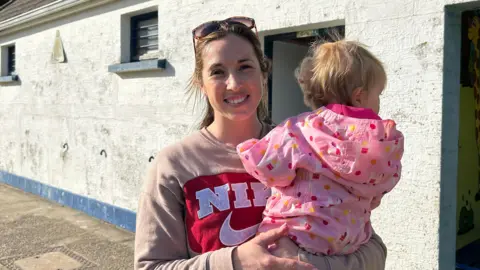 Claudia is wearing a brown Nike Jumper with sunglasses and is holding her baby who is wearing a pink jumpsuit