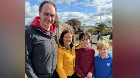 Keir Mackenzie/BBC John O’Driscoll, Sarah O’Driscoll and their children Fin and Zac at Mr Mulligan's Dino Golf in Tonbridge, Kent. All four are holding golf clubs. John and Sarah are wearing puffer jackets, while Fin and Zac are wearing hoodies. The golf course can be seen in the background.