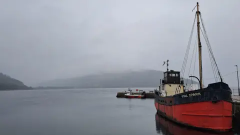 Charlotte McAllister A red-and-black vintage boat named “Vital Spark” is moored beside a calm harbour on a misty day. Low clouds and fog partially obscure the hills in the background. A smaller boat is tied up at a nearby pier, while the still water reflects the larger vessel. 