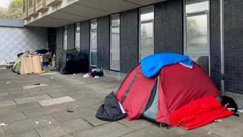 A red tent with a blue sleeping bag draped over the top with three more tents pitched on paving stones in the background 