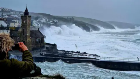 A person with blonde hair takes a picture of the bay around Porthleven in Cornwall during Storm Kathleen. Three seagulls are flying around while waves crash into the coastline in the distance.