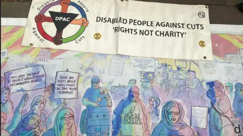A close up of an ornate banner showing people on a picket line. At the top of the banner it reads: "Disabled people against cuts. Rights not charity."
