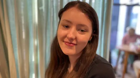 Laura Foster/BBC Lauren is a young woman with long brown hair which she is wearing down. Her hair is tucked behind her ears and she's wearing a black top and looking at the camera smiling. Behind her is a pale green curtain.