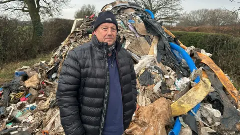 Councillor Phil Whitehouse, standing next to a large pile of waste material. He is wearing a black jacket over a blue jumper as well as a black woollen hat.