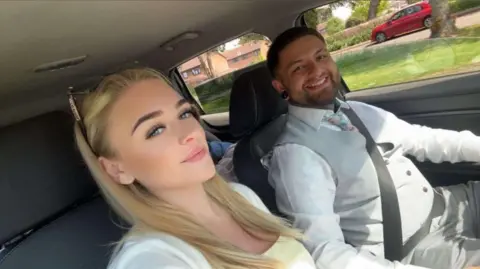 Brad Pruden A woman and a man smile towards the camera while sat inside a car, with the woman wearing a yellow dress and man wearing a waistcoat and smart shirt