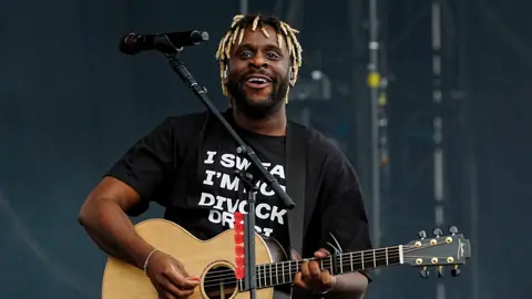 Myles Smith performs onstage, with an acoustic guitar and a black T-shirt on.
