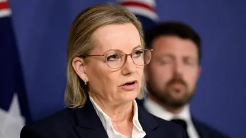 Sussan Ley, wearing glasses and a black jacket, speaks, while a man with a beard stand behind, not in focus. 