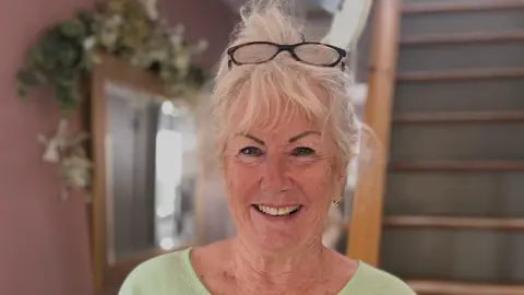 The image shows a woman standing indoors near the bottom of a wooden staircase, wearing a light green top with a pair of glasses resting on her head. Behind the woman is a pale pink wall with a rectangular mirror and decorative greenery.