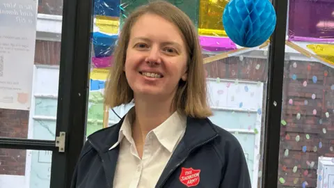 A woman with a blonde bob haircut wearing a white shirt and dark jacket which has a red badge on with The Salvation Army in white letters. She is smiling at the camera and behind her are colourful decorations and glass doors. 