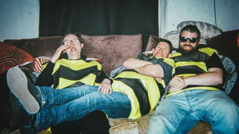Ben G. Brown Three people, wearing matching yellow and black stripy t-shirts and blue jeans, lounge on a large sofa. 