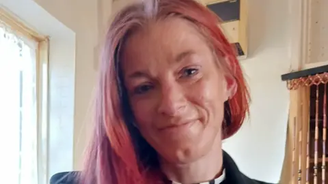 A woman with pink long hair smiling. She is standing next to a wind