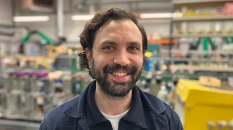 Sam Read/BBC A man wearing a blue jacket and white T-shirt who is smiling looking into the camera. He has dark brown hair and a short beard and moustache. He is standing in a factory.