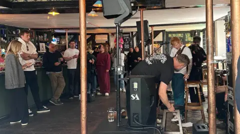 Richard Knights/BBC Ed Sheeran with band crew setting up for a perfomance in a pub. people stand about the bar drinking and looking on.