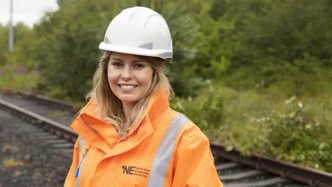 Kim McGuinness is smiling at the camera in a white helmet and orange high visibility protective clothing. She is standing at the currently disused Leamside Line, with the overgrown tracks behind her.