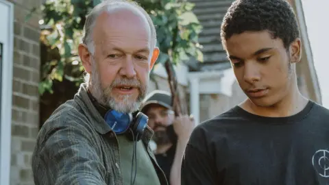 Jessica Sansom Two people talking. The man on the left is older with a grey beard and short hair. He is slightly bald. He has headphones around his neck. he is explaining something to a young male actor who is listening intently to him.