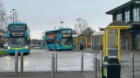 Two blue buses are stopped at bus stops outside Morrisons supermarket. A third bus is in the distance driving down a residential road.
