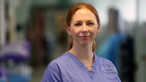 Moondance Cancer Initiative Jody is wearing blue theatre scrubs and there is a blurred hospital corridor in the background.