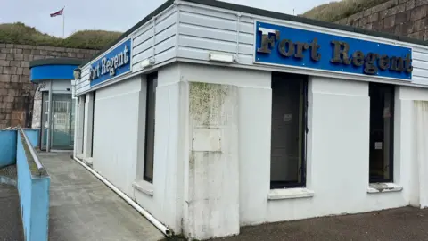 A single storey white building with blue signs with silver letters reading Fort Regent on both sides. The building has windows with closed blinds and has signs of damp. There is a ramp with a blue hand rail leading to a rounded entrance with glass doors. Surrounding the building is a brick wall behind which are two hills which have a UK flag flying between them.