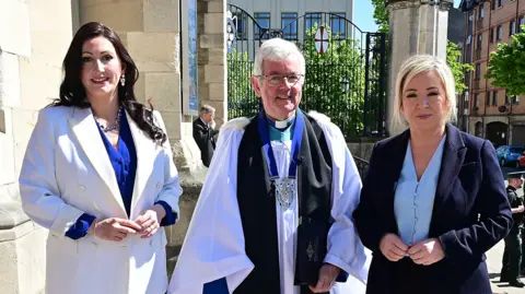 Emma Little-Pengelly stands with the Dean of Belfast Rev Stephen Forde and Michelle O'Neill on the steps outside Belfast Cathedral. Little-Pengelly stands on the left wearing a blue dress and white blazer, and has long, dark hair. In the middle is Rev Forde, who is wearing a white robe with a black long scarf. He has short white hair and is wearing silver round glasses. On the right is O'Neill, who is wearing a navy suit and light blue button up shirt and has her blonde hair tied back.