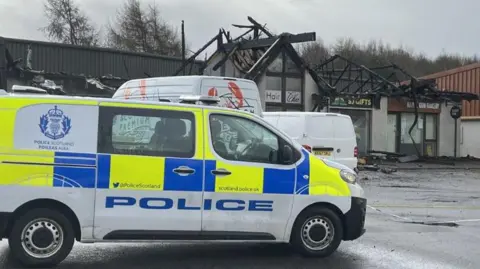 BBC A police van sits in front of a burnt out row of shops