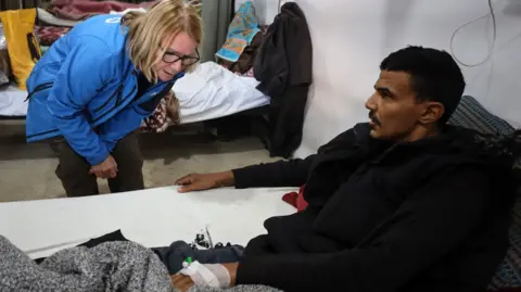 UK-Med A woman with blonde hair and glasses, wearing a blue jacket, leans in to look at the canular on the back of a man's hand. He is laying on a bed and wearing back clothing