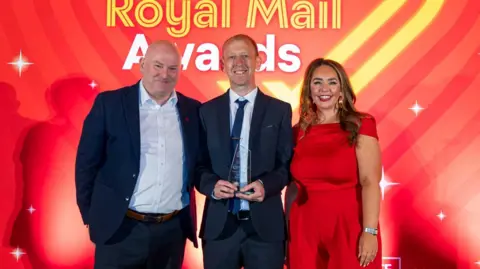 Royal Mail Richard Kesterton stood in front of a red background holding a glass award. He is wearing a dark blue suit with white shirt and blue tie, while stood in between another man wearing a dark suit and white shirt open at the top, and a woman in a red dress. Richard has short brown hair and is smiling at the camera.