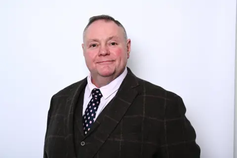 Labour Council leader Stephen Simkins, wearing a brown check suit and tie standing against a white background