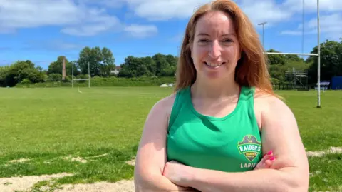 BBC A smiling woman with her arms crossed, wearing a green Guernsey Raiders Ladies training vest. She stands in front of a rugby pitch.