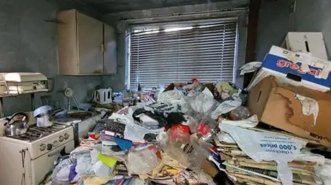 A kitchen with a hob, shelves and kettle in the background with piles of paperwork and litter. A broken window blind covered in dirt hangs in the window. 