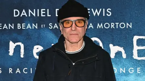 Daniel Day-Lewis staring at the camera. He is wearing sunglasses, a black hat and black jacket
