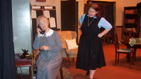supplied An elderly woman sits on stage dressed in a floor length grey dress, answering an old fashioned black telephone