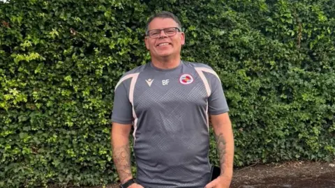 A picture of Brendon Flanagan, who is wearing a grey Reading FC shirt with "BF" on the front of it. He is smiling and has greying dark hair and is wearing black-rimmed glasses.