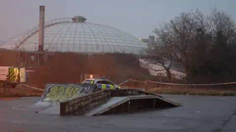A skate park at dusk. The equipment is heavily graffitied. Police tape cordons off the scene. A police car and a police van is visible. The dome of the Oasis centre sits in the background. 