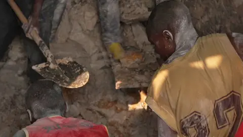 Two miners with their backs to the camera bend over as they dig in a pit at Rubaya mine in eastern DR Congo. The one in the left wears a red tank top and his colleague on the right wears a yellow sports shirt with numbers printed on the back. In the background the metal spade of another miner can be seen.