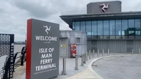 The exterior of the ferry terminal in Liverpool, which has a sign that reads Welcome to the Isle of Man Ferry Terminal. There is a three legs of man sign on the building, which has large windows and is grey. 