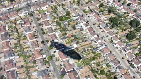 Looking down from a blimp onto residential streets and back gardens, with the aircraft's shadow clearly visible on the ground.