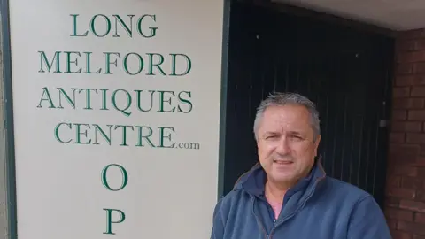 Supplied A head and shoulders image of Geoffrey McGregor. He is wearing a navy blue fleece and standing in front of a white and green sign which reads Long Melford Antiques Centre. He is smiling and looking into the camera.