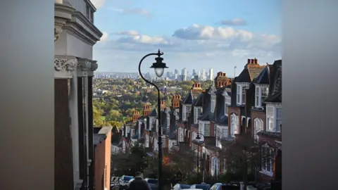 Getty Images A steep hilly suburban street lined with red brick Victorian houses. In the background is central London and a blue sky with white clouds. 