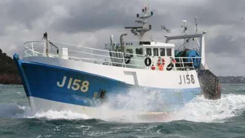 States of Jersey Police A white and blue fishing boat with J158 on the left side, in the ocean sailing. Land can been seen afar in the background.