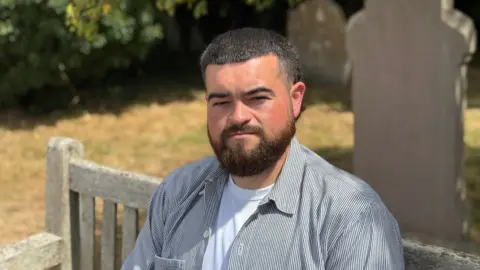 Sam Wilmot looks straight at the camera sitting on a wooden bench, with gravestones in the background.