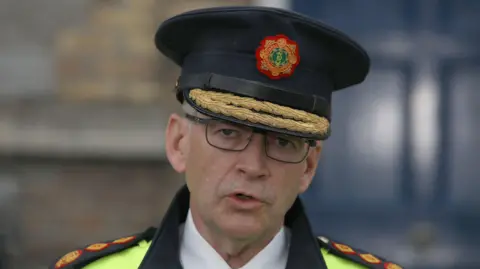 PA Media A close-up image of Garda Commissioner Drew Harris. He is an older man who is wearing a black, flat top hat with the garda logo in the centre and two gold ropes on the brim. He is mid-speech and is wearing a pair of grey metal glasses. He is also wearing a reflective police jacket and a white collared shirt.